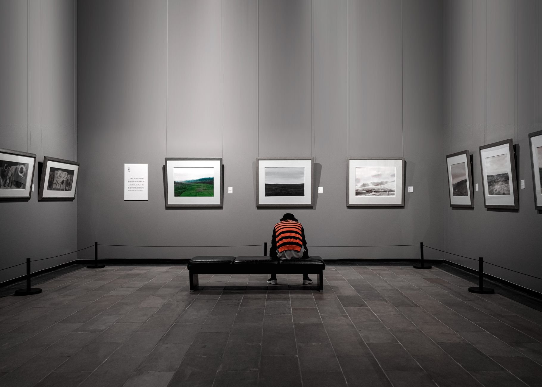 A solitary figure sitting in a contemporary art gallery surrounded by framed artworks.